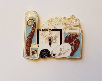 Puede incluir: Pin esmaltado que representa una colección de especímenes conservados. Incluye un frasco con un tentáculo de pulpo, una seta, una mariposa, un corazón, una calavera, un esqueleto de camaleón y una concha de caracol. El pin tiene contornos dorados.