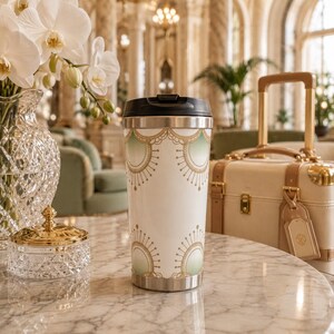 May include: A white travel tumbler with a black lid featuring a repeating pattern of gold scalloped arches and sunburst motifs with soft green gradient accents. The cylindrical container has a metallic base and a smooth, glossy finish.