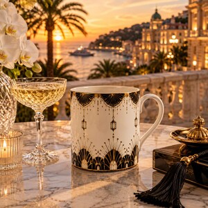 May include: A white ceramic mug featuring a black and gold Art Deco-inspired fan pattern with hanging lantern motifs. The mug has a curved handle and sits on a marble surface next to a crystal coupe glass and a black tassel accessory.