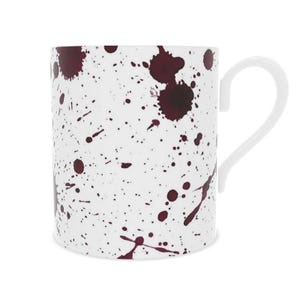 May include: White ceramic mug featuring a burgundy splatter design. The mug has a curved white handle and a glossy finish. The splatter pattern varies in size and density across the surface.