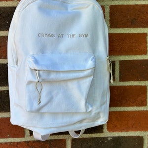 Puede incluir: Mochila blanca con un bolsillo frontal y el texto bordado "CRYING AT THE GYM". La mochila tiene un asa superior, correas ajustables para los hombros y un compartimento principal con cremallera. La bolsa está hecha de tela texturizada.
