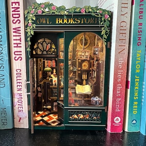 May include: A miniature wooden model of a bookstore with a green exterior and a sign that reads "Owl Bookstore". The storefront has a glass window with a view of the interior, which includes bookshelves, a desk, and a clock. The model is sitting on a bookshelf with other books.