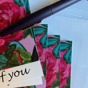 May include: A stack of blank greeting cards with a floral design featuring pink roses and green leaves. The text "Thinking of you" is printed on the front of the top card. A purple pen is lying on top of the cards.