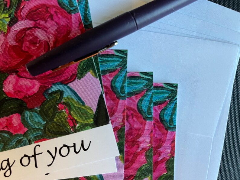 May include: A stack of blank greeting cards with a floral design featuring pink roses and green leaves. The text "Thinking of you" is printed on the front of the top card. A purple pen is lying on top of the cards.