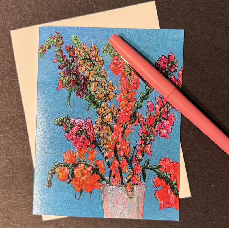 Peut inclure: Carte de v&oelig;ux avec une peinture florale de fleurs rouges, oranges et roses dans un vase blanc sur fond bleu clair. Un stylo rose est pos&eacute; en diagonale sur la carte. La carte est sur une surface marron fonc&eacute;.