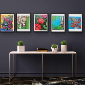 Op de afbeelding: Vijf ingelijste kunstafdrukken met kleurrijke bloemen- en dierenillustraties. De prints worden tentoongesteld boven een witte marmeren console met decoratieve planten. Het kunstwerk bevat een hond, een konijn en diverse bloemen.