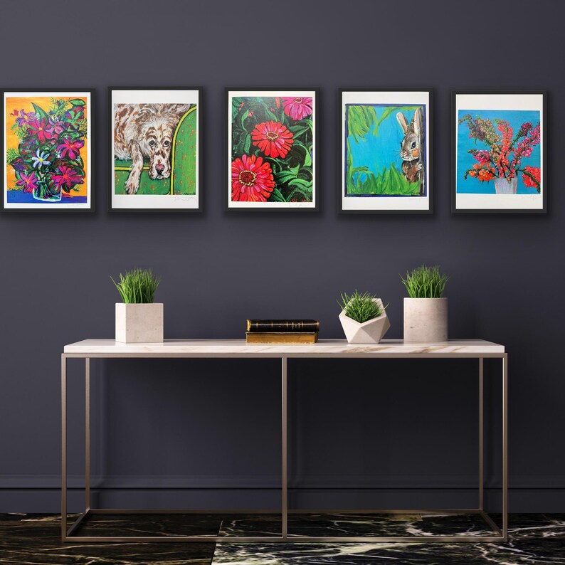 Op de afbeelding: Vijf ingelijste kunstafdrukken met kleurrijke bloemen- en dierenillustraties. De prints worden tentoongesteld boven een witte marmeren console met decoratieve planten. Het kunstwerk bevat een hond, een konijn en diverse bloemen.