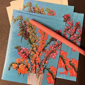 Peut inclure: Cartes de v&oelig;ux avec un motif floral. Les cartes pr&eacute;sentent un bouquet de fleurs orange, roses et jaunes dans un vase blanc sur un fond bleu clair textur&eacute;. Un stylo rose est pos&eacute; dessus.