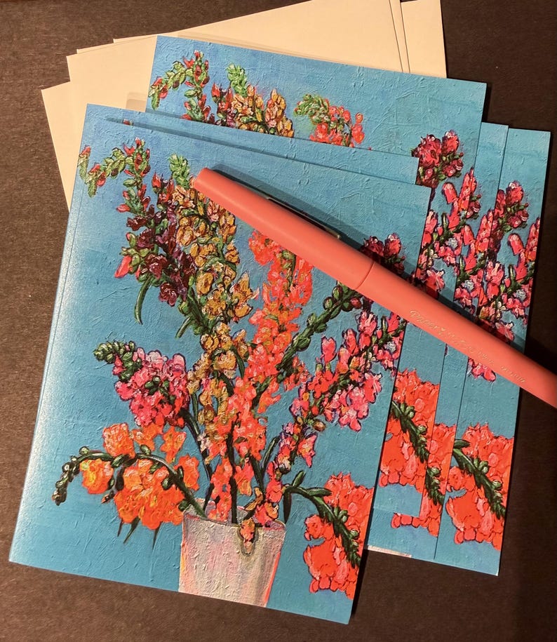 Peut inclure: Cartes de v&oelig;ux avec un motif floral. Les cartes pr&eacute;sentent un bouquet de fleurs orange, roses et jaunes dans un vase blanc sur un fond bleu clair textur&eacute;. Un stylo rose est pos&eacute; dessus.