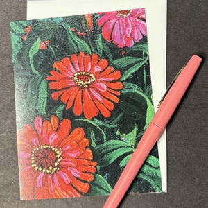 Op de afbeelding: Wenskaart met een schilderij van rode en roze zinnia's en groene bladeren. Een roze pen ligt naast de kaart en een witte envelop. Het kunstwerk heeft een textuur, schilderachtige stijl.