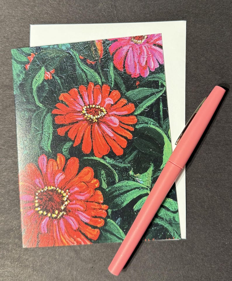Op de afbeelding: Wenskaart met een schilderij van rode en roze zinnia's en groene bladeren. Een roze pen ligt naast de kaart en een witte envelop. Het kunstwerk heeft een textuur, schilderachtige stijl.