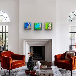 Peut inclure: Trois peintures carr&eacute;es de lapins sont accroch&eacute;es &agrave; un mur blanc. Chaque peinture pr&eacute;sente une sc&egrave;ne de lapin diff&eacute;rente avec des couleurs vives. L'&oelig;uvre est expos&eacute;e au-dessus d'une chemin&eacute;e, avec deux fauteuils orange au premier plan.