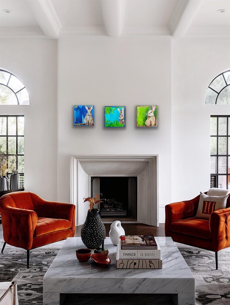Peut inclure: Trois peintures carr&eacute;es de lapins sont accroch&eacute;es &agrave; un mur blanc. Chaque peinture pr&eacute;sente une sc&egrave;ne de lapin diff&eacute;rente avec des couleurs vives. L'&oelig;uvre est expos&eacute;e au-dessus d'une chemin&eacute;e, avec deux fauteuils orange au premier plan.