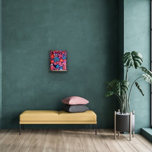 May include: A framed abstract painting with a vibrant mix of red, pink, and blue flowers on a dark blue background. The painting is hanging on a teal wall in a modern living room setting.