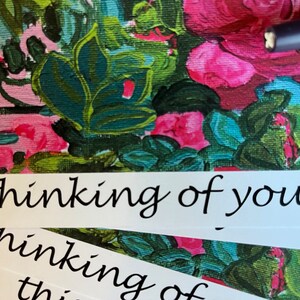 May include: A white card with the text "thinking of you" written in black cursive script. The card is on top of a floral patterned surface.