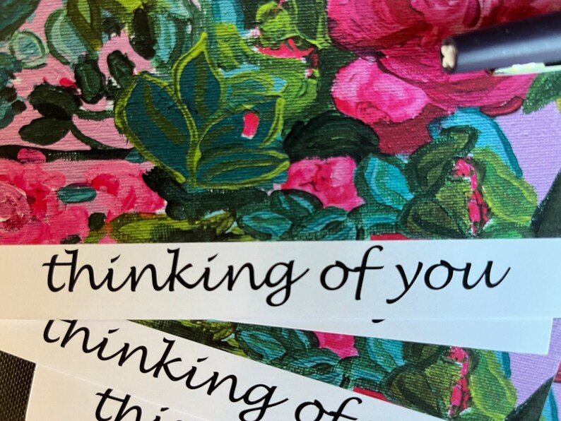 May include: A white card with the text "thinking of you" written in black cursive script. The card is on top of a floral patterned surface.