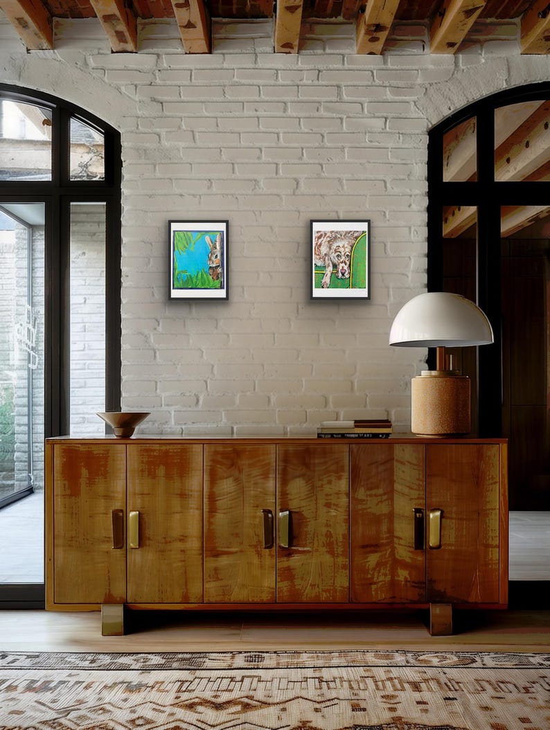 Op de afbeelding: Twee ingelijste kunstafdrukken hangen aan een witte bakstenen muur. E&eacute;n toont een konijn tegen een blauwe achtergrond, de andere een hond op groen. Daaronder een houten kast met gouden handgrepen en een lamp met een beige voet.