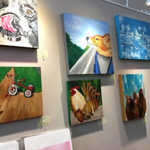 Op de afbeelding: Een galeriewand met verschillende schilderijen met daarop een hond, een haan, kippen, een driewieler en een hond. De schilderijen zijn ingelijst en hangen aan een witte muur.