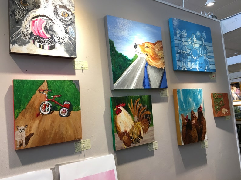 Op de afbeelding: Een galeriewand met verschillende schilderijen met daarop een hond, een haan, kippen, een driewieler en een hond. De schilderijen zijn ingelijst en hangen aan een witte muur.