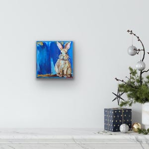 Peut inclure: Tableau carr&eacute; repr&eacute;sentant un lapin assis, peint en marron et blanc sur un fond bleu. L'&oelig;uvre d'art pr&eacute;sente une texture avec des coups de pinceau visibles. Le tableau est accroch&eacute; sur un mur blanc.