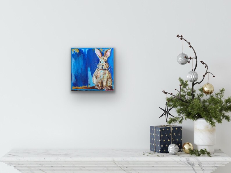 Peut inclure: Tableau carr&eacute; repr&eacute;sentant un lapin assis, peint en marron et blanc sur un fond bleu. L'&oelig;uvre d'art pr&eacute;sente une texture avec des coups de pinceau visibles. Le tableau est accroch&eacute; sur un mur blanc.