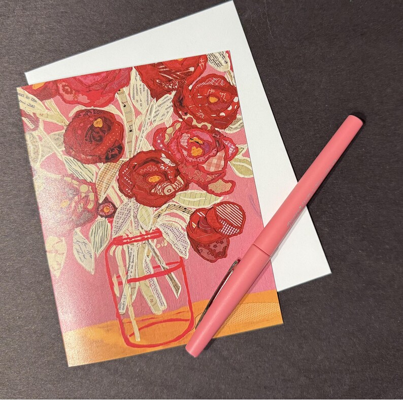 May include: Greeting card featuring a floral design with red roses in a clear jar, set against a pink background. A pink pen rests beside the card and a white envelope. The card's design includes a collage of textures and patterns.