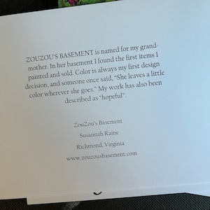 May include: A white card with black text describing the artist's inspiration for their art. The text reads "ZOUZOU'S BASEMENT is named for my grandmother. In her basement I found the first items I painted and sold. Color is always my first design decision, and someone once said, "She leaves a little color wherever she goes." My work has also been described as "hopeful."". The card also includes the artist's name, location, and website: "ZouZou's Basement, Susannah Raine, Richmond, Virginia, www.zouzousbasement.com".