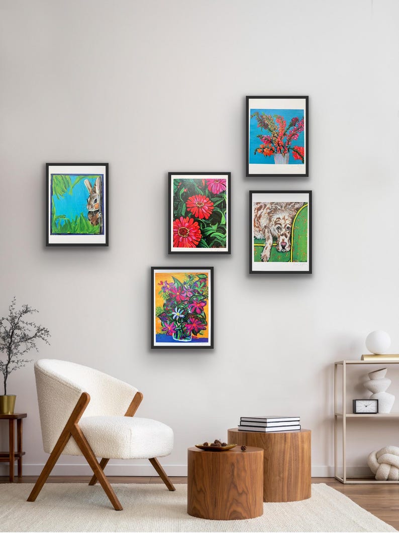 Op de afbeelding: Vijf ingelijste kunstdrukken tentoongesteld op een witte muur. De prints tonen kleurrijke bloemstukken, een konijn en een hond. De frames zijn zwart en de kunstwerken hebben levendige kleuren. De kamer bevat een witte stoel en houten bijzettafels.