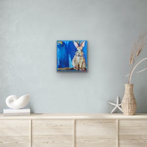 Peut inclure: Tableau carr&eacute; repr&eacute;sentant un lapin assis aux longues oreilles, peint dans des tons de beige et de blanc. Le lapin se d&eacute;tache sur un fond bleu vibrant avec des coups de pinceau. L'&oelig;uvre est encadr&eacute;e d'une bordure dor&eacute;e.