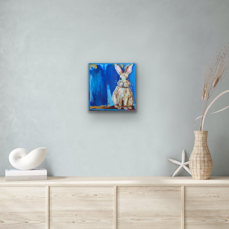 Peut inclure: Tableau carr&eacute; repr&eacute;sentant un lapin assis aux longues oreilles, peint dans des tons de beige et de blanc. Le lapin se d&eacute;tache sur un fond bleu vibrant avec des coups de pinceau. L'&oelig;uvre est encadr&eacute;e d'une bordure dor&eacute;e.