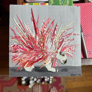 Pu&ograve; includere: Un'opera d'arte quadrata con un collage di porcospino. Gli aculei del porcospino sono fatti di carta a motivi rossi e bianchi. L'animale ha una faccia e zampe bianche, su sfondo grigio. La firma dell'artista, "SUSANNAH", &egrave; visibile.