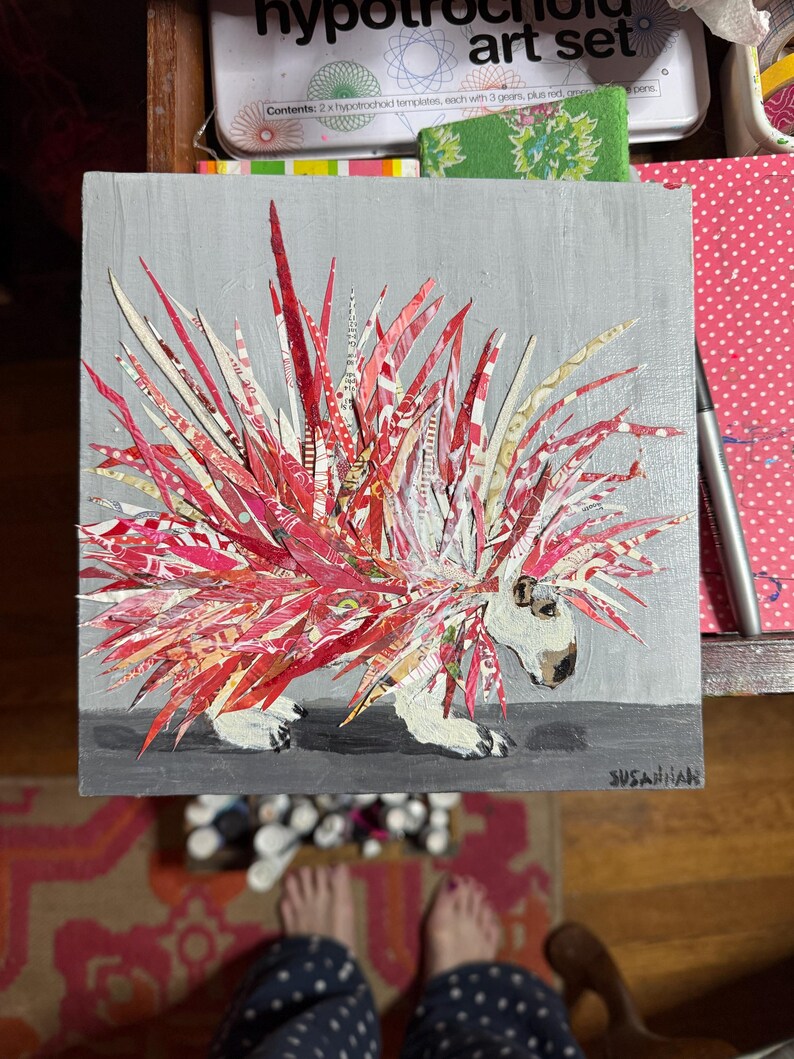 Pu&ograve; includere: Un'opera d'arte quadrata con un collage di porcospino. Gli aculei del porcospino sono fatti di carta a motivi rossi e bianchi. L'animale ha una faccia e zampe bianche, su sfondo grigio. La firma dell'artista, "SUSANNAH", &egrave; visibile.