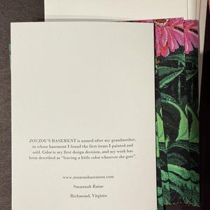 Op de afbeelding: Een stapel witte kaarten met tekst van "ZOUZOU'S BASEMENT" en een bloemenontwerp. De tekst bevat de website, de naam van de kunstenaar en de locatie. Het bloemenontwerp toont roze bloemen en groene bladeren.