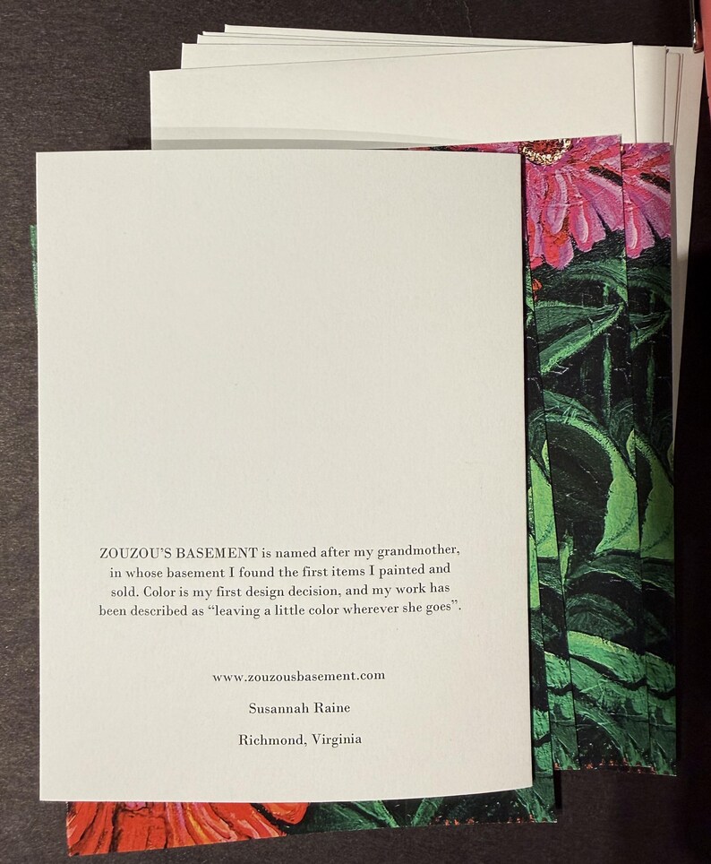 Op de afbeelding: Een stapel witte kaarten met tekst van "ZOUZOU'S BASEMENT" en een bloemenontwerp. De tekst bevat de website, de naam van de kunstenaar en de locatie. Het bloemenontwerp toont roze bloemen en groene bladeren.