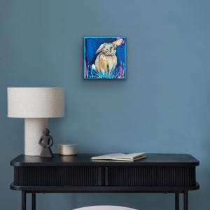 May include: Square painting of a dog with floppy ears, set against a blue background. The dog is painted in shades of tan and brown, with pink and blue accents around it. The artwork is displayed on a blue wall.