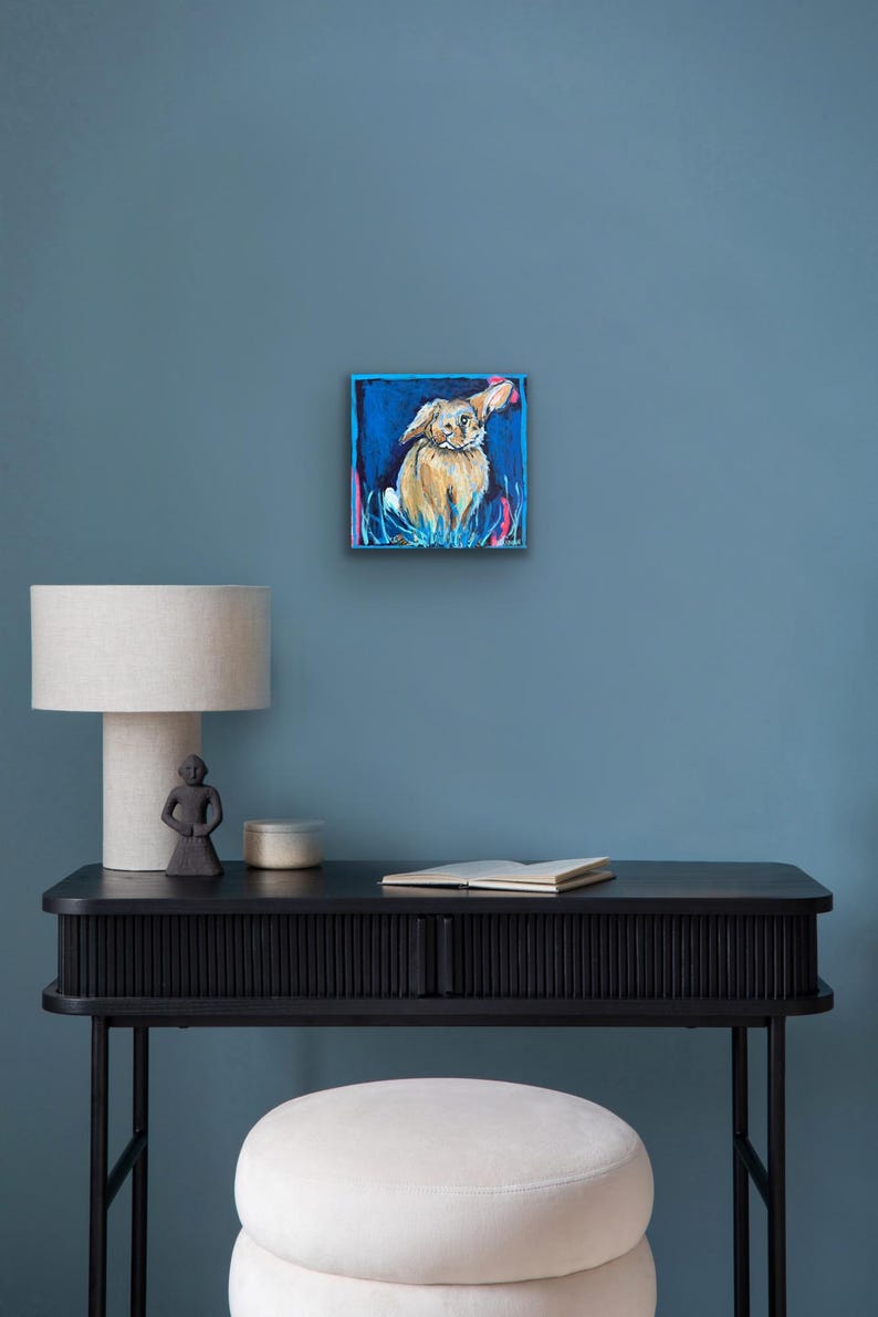 May include: Square painting of a dog with floppy ears, set against a blue background. The dog is painted in shades of tan and brown, with pink and blue accents around it. The artwork is displayed on a blue wall.