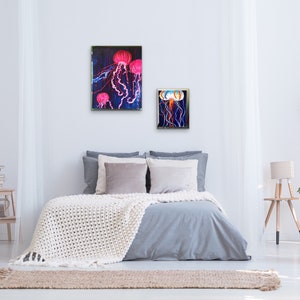 May include: Two framed paintings of jellyfish in a bedroom setting. The larger painting features three pink jellyfish with long tentacles against a dark blue background. The smaller painting features one jellyfish with a white and blue body and long tentacles against a dark blue background.