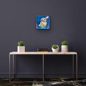May include: Square painting of a rabbit in a blue frame. The rabbit is tan with a white chest and is surrounded by blue and pink brushstrokes. The artwork is displayed on a white marble table with decorative plants.
