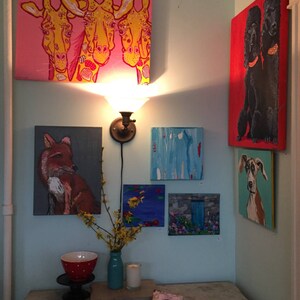 May include: A collection of framed paintings featuring various animal subjects, including giraffes, a fox, a dog, and two poodles. The paintings are displayed on a light blue wall with a small table and a vase of yellow flowers in the foreground.