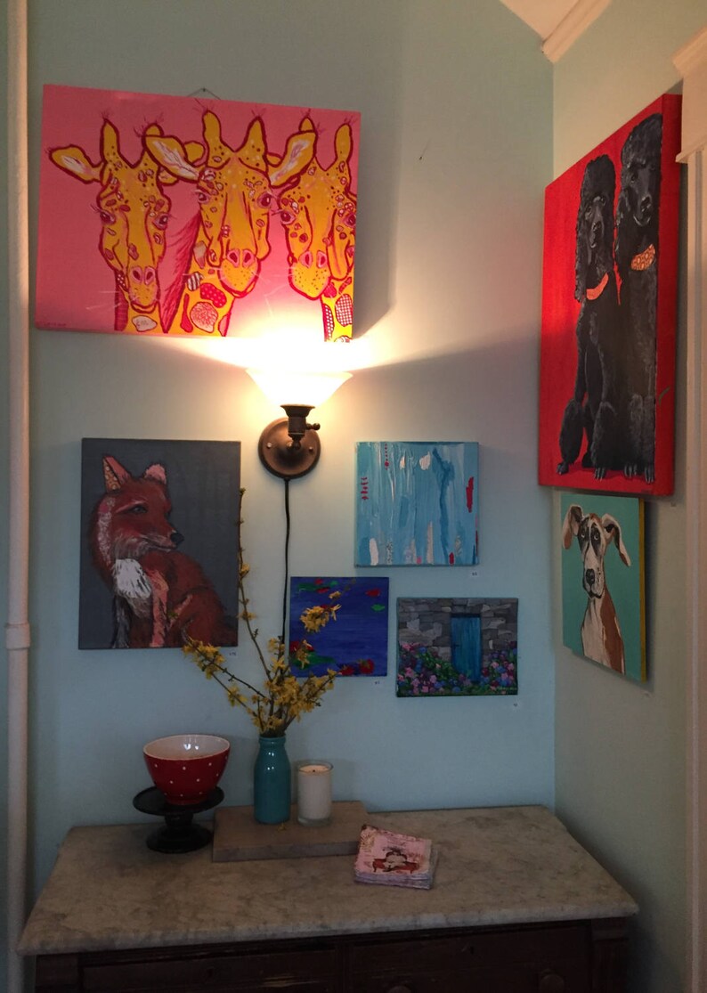 May include: A collection of framed paintings featuring various animal subjects, including giraffes, a fox, a dog, and two poodles. The paintings are displayed on a light blue wall with a small table and a vase of yellow flowers in the foreground.
