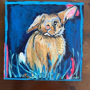 May include: A square painting of a brown rabbit with long ears, set against a deep blue background. The rabbit is painted with textured brushstrokes, and the ears have pink accents. The artwork includes blue and pink vertical strokes and the artist's signature.