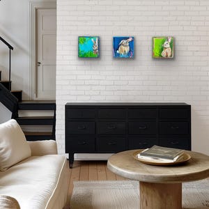May include: Three square framed paintings of rabbits hang on a white brick wall. Each painting features a different rabbit in a colorful, abstract setting. The room includes a black dresser, a white sofa, and a wooden coffee table.