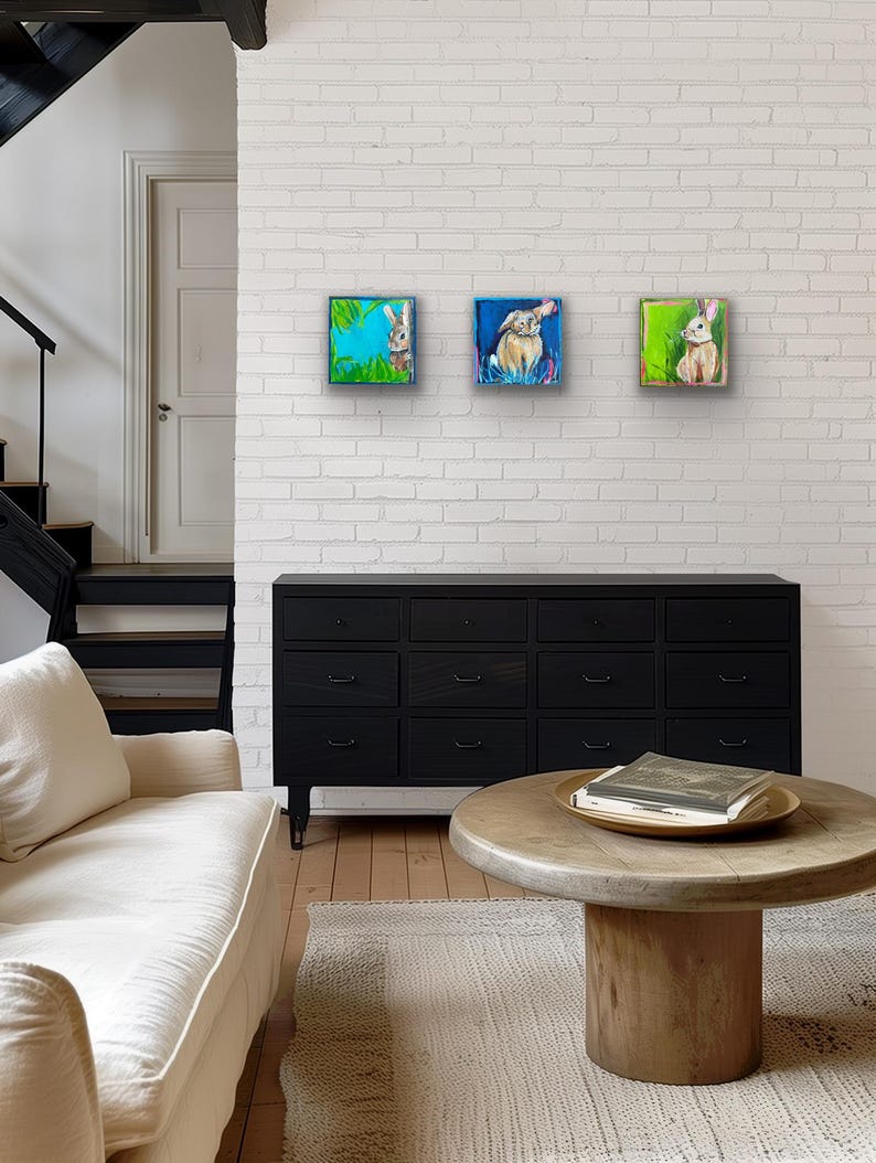 May include: Three square framed paintings of rabbits hang on a white brick wall. Each painting features a different rabbit in a colorful, abstract setting. The room includes a black dresser, a white sofa, and a wooden coffee table.