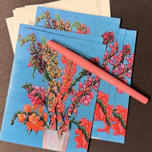 Peut inclure: Un ensemble de cartes de v&oelig;ux florales avec un fond bleu clair et un stylo rose. Les cartes pr&eacute;sentent une peinture color&eacute;e de fleurs dans un vase. Des enveloppes blanches sont empil&eacute;es en arri&egrave;re-plan.