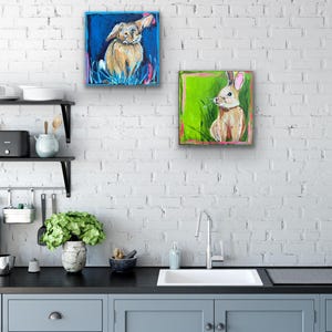 May include: Two square paintings of rabbits. One painting features a tan rabbit with a blue background. The other painting shows a rabbit with a green background. Both paintings have a colorful, whimsical style.