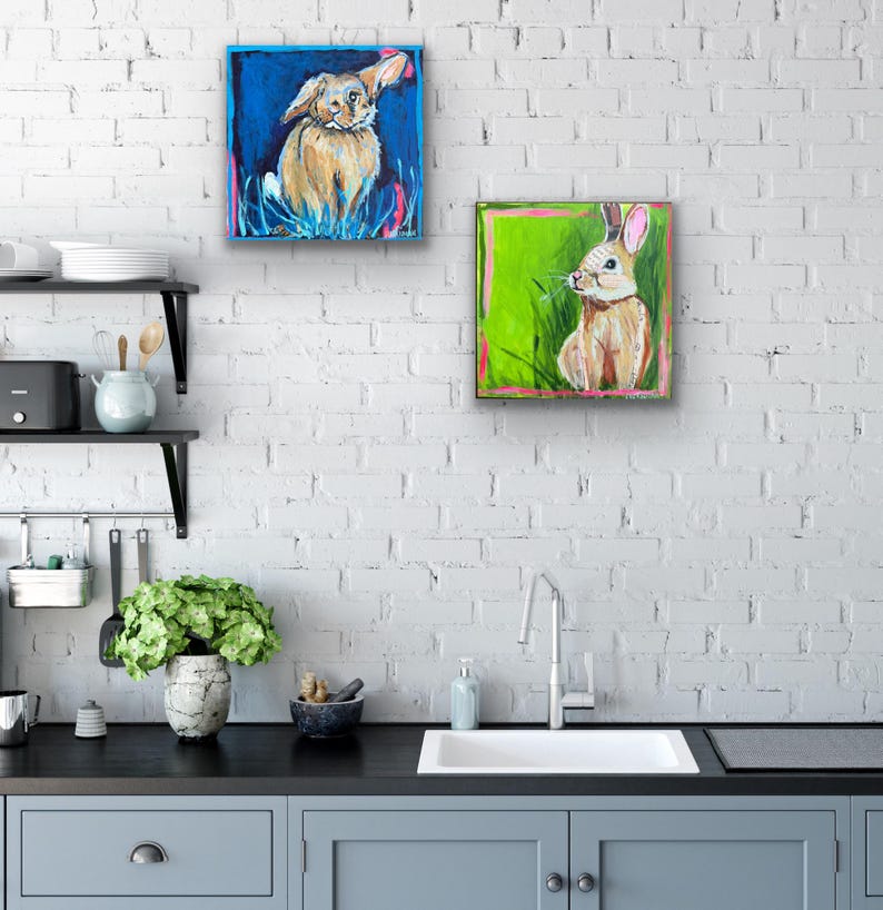 May include: Two square paintings of rabbits. One painting features a tan rabbit with a blue background. The other painting shows a rabbit with a green background. Both paintings have a colorful, whimsical style.