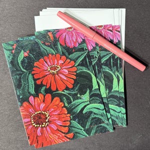 Op de afbeelding: Een stapel bloemrijke wenskaarten met levendige rode en roze zinnia bloemen en groene bladeren. Een roze pen ligt bovenop de kaarten en witte enveloppen.