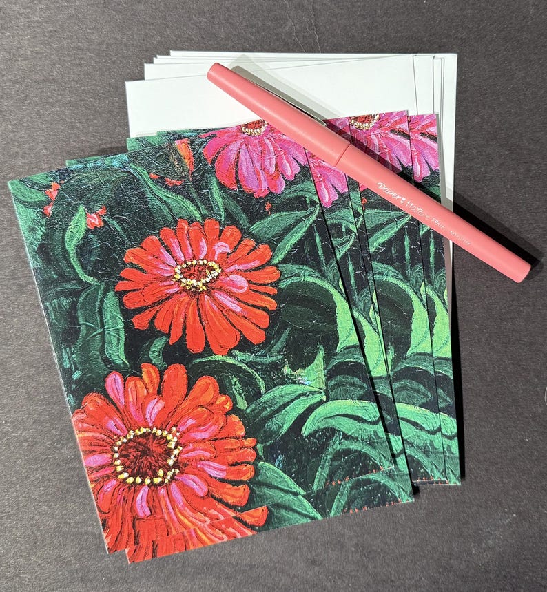 Op de afbeelding: Een stapel bloemrijke wenskaarten met levendige rode en roze zinnia bloemen en groene bladeren. Een roze pen ligt bovenop de kaarten en witte enveloppen.
