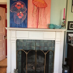 May include: Two paintings of jellyfish, one with a blue background and a pink jellyfish, the other with a pink background and a red jellyfish. The paintings are hanging above a white fireplace with a black grate.