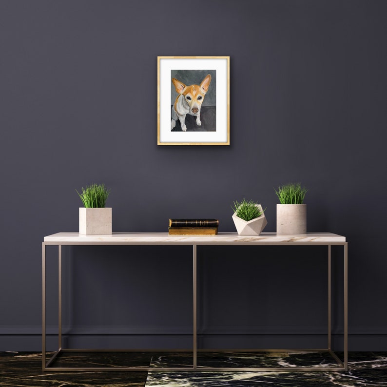 May include: A framed watercolor painting of a small, white and brown dog with floppy ears, looking directly at the viewer. The dog is sitting on a gray background. The frame is a light wood color.